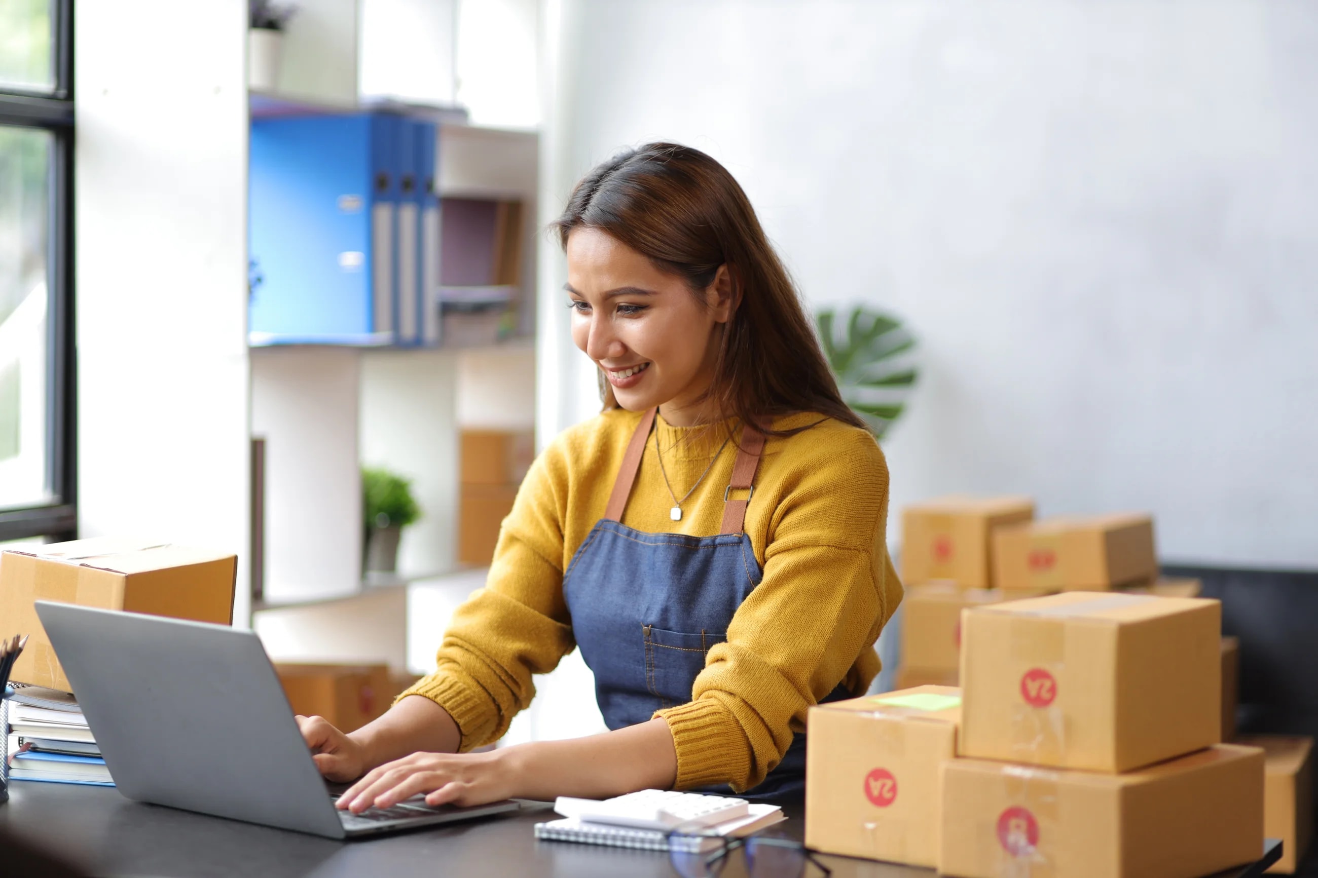Emprendedora sonriendo mientras gestiona pedidos en su laptop, con cajas de envío de su tienda en línea a un lado